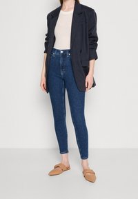 Tmavě modré vysoké skinny džíny, spárované s obepnutým béžovým topem a volným tmavým sakem. Součástí outfitu jsou béžové skluzavkové sandály.
