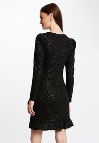 Robe noire à manches longues, motif floral texturé, corsage ajusté et ourlet à volants évasé. Tissu lisse avec un léger éclat.
