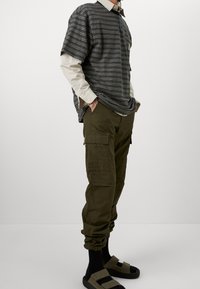 Pasiasta, czarno-szara, oversize'owa koszulka nałożona na kremową bluzkę z długim rękawem, zestawiona z oliwkowymi spodniami cargo i czarnymi sandałami.