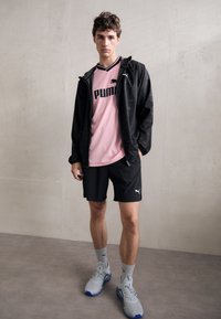 Rosa sportsdrakt med svart logo, svart glidelåsjakke, svarte shorts, grå sko med blå detaljer, stående på en nøytral gulv.