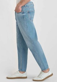 Hombre vestido con jeans de mezclilla azul claro y zapatillas blancas, de pie con una mano en el bolsillo frente a un fondo liso.