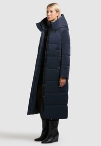 Lang marineblå pufferjakke med høj krave, vandret syninger og sidelommer med lynlås. Stoffet er glat og skinnende; parret med sorte fodtøj.