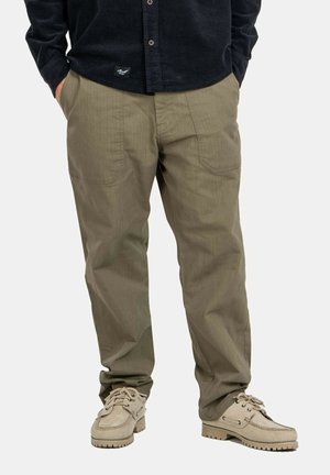 Mann trägt olivgrüne Hose mit aufgesetzten Fronttaschen, beige Schnürschuhe und ein schwarzes Hemd mit Knöpfen, die Hände in den Taschen.