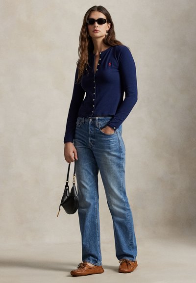 Sieviete valkā tumši zilu pogātu kardiganu, zilas džinsas, brūnas mokasīnus, melnas saulesbrilles, turot melnu rokassomiņu pret neitrālu fonu.