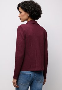 Pull texturé bordeaux avec col, manches longues et coupe carrée. Porté avec un jean en denim bleu, présentant une couture minimale sur les poches.