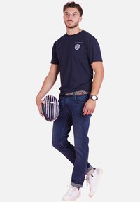 T-shirt marine avec logo, jean en denim foncé et baskets blanches. Tient un ballon de rugby rayé, présenté avec une ceinture à motif.