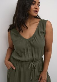 Olive green sleeveless jumpsuit made of lightweight, textured fabric. Features a v-neckline, gathered detail, and adjustable drawstring waist.