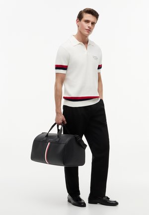 Junger Mann trägt weißes Poloshirt mit roten und marineblauen Streifen, schwarze Hose, schwarze Schuhe, hält schwarze Reisetasche mit roten, weißen und marineblauen Streifen.