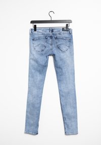 Vaqueros ajustados de mezclilla azul claro con textura suave, cinco bolsillos y una pequeña etiqueta de marca en la parte trasera. Cuelgan de una percha negra.