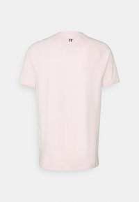 Camiseta rosa claro de manga corta en algodón suave, con un escote redondo y un pequeño logo negro en la parte superior de la espalda. Textura suave.
