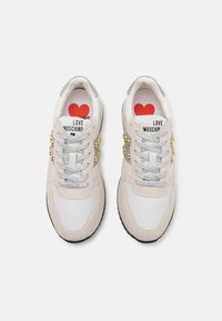 Zapatillas deportivas blancas y beige con una suela gruesa, acentos de ante texturizado, cordones grises y tachuelas doradas. Presentan un logotipo de corazón rojo en el interior.