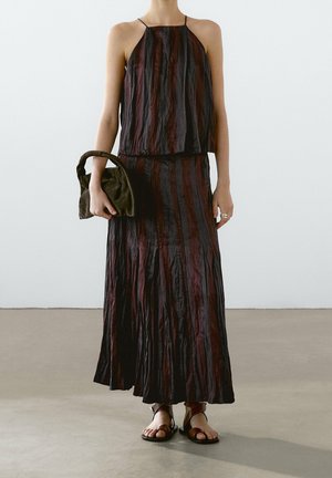 Woman wearing sleeveless, striped, crinkled maxi dress in dark red and black, holding a soft, olive green handbag and wearing brown sandals.