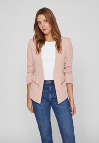 Blazer rosa claro con un diseño ajustado, mangas largas y bolsillos con cremallera, combinado con una blusa blanca y jeans azul oscuro.