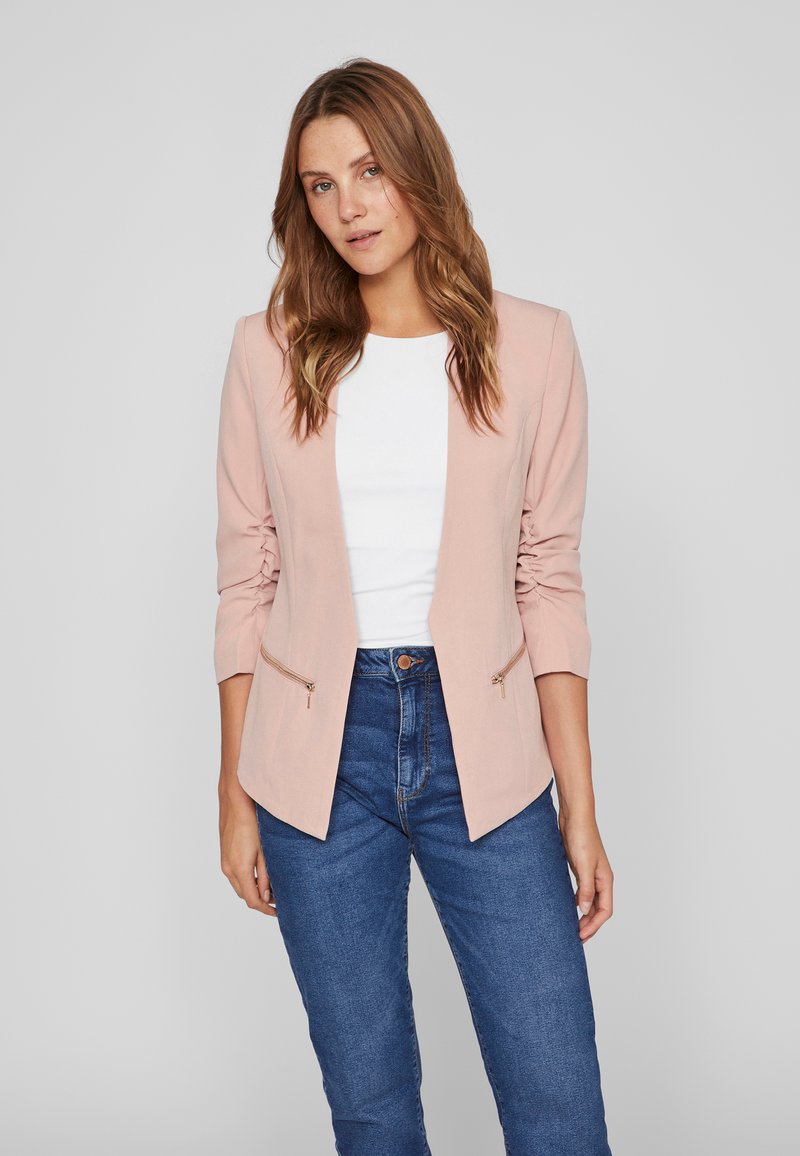 Blazer rosa claro con un diseño ajustado, mangas largas y bolsillos con cremallera, combinado con una blusa blanca y jeans azul oscuro.