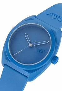 Kék szilikon óra kerek számlappal, minimalista dizájn, díszítmények nélküli óra és perc mutatók. Logó az alsó bal sarokban.