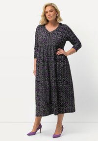 Schwarzes Blumenkleid mit lila Akzenten, V-Ausschnitt und dreiviertel Ärmeln. Das Model kombinierte es mit lila High Heels. Länge bis zur Mitte der Waden.