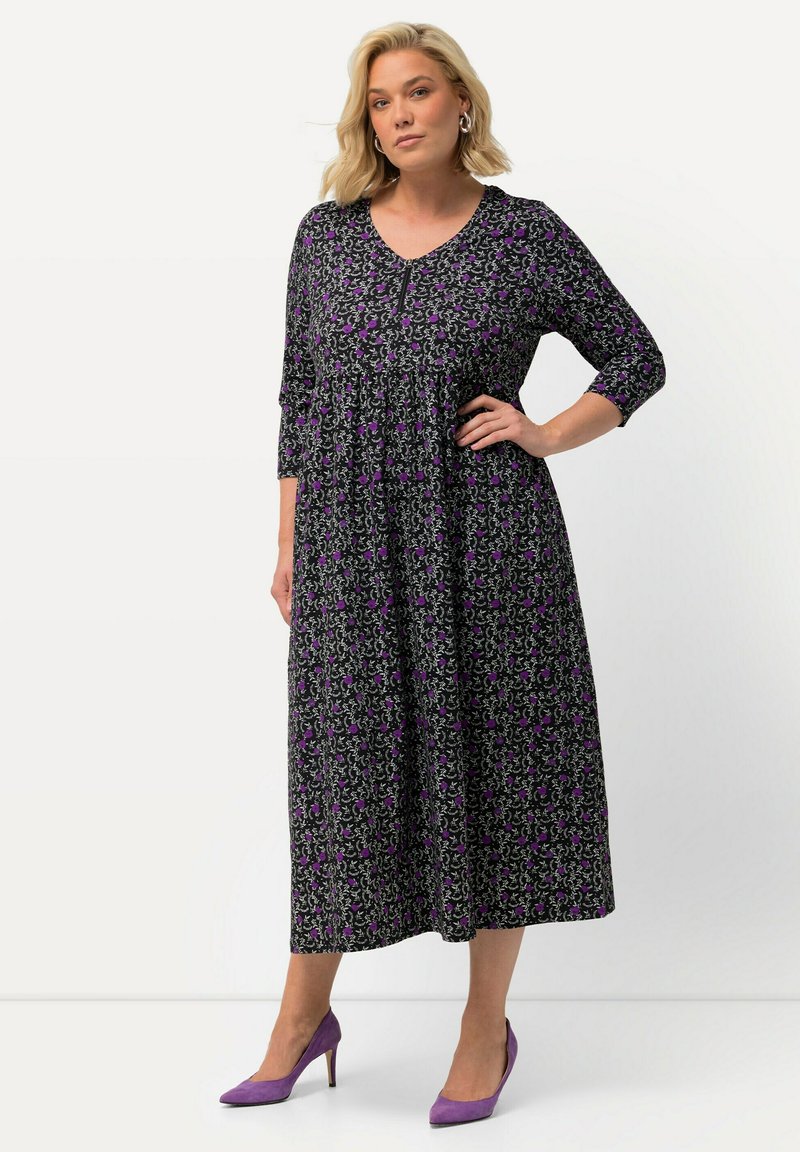 Schwarzes Blumenkleid mit lila Akzenten, V-Ausschnitt und dreiviertel Ärmeln. Das Model kombinierte es mit lila High Heels. Länge bis zur Mitte der Waden.