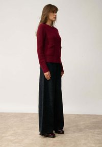 Bordeaux geribbelde gebreide trui met twee voorkeurszakken, gecombineerd met brede zwarte corduroy broek. Model staat tegen een neutrale achtergrond.