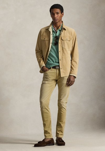 Μπεζ σουέντ μπουφάν πάνω από πράσινο πουλόβερ, σετ με ανοιχτό κίτρινο παντελόνι και καφέ μοκασίνια. Σημαντικά αξεσουάρ είναι τα γυαλιά ηλίου και ένα φουλάρι με σχέδια.