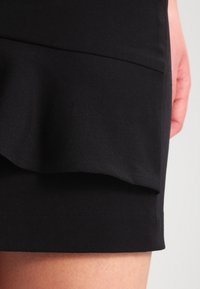 Gonna nera con orlo arricciato, texture liscia e design aderente. Il tessuto sembra resistente con una leggera lucentezza.