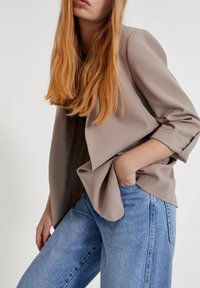 Jasnobeżowy żakiet z podwiniętymi rękawami, zwisający materiał, noszony na ciemnej bluzce, zestawiony z wysokim stanem niebieskich dżinsów, widoczna przednia kieszeń.