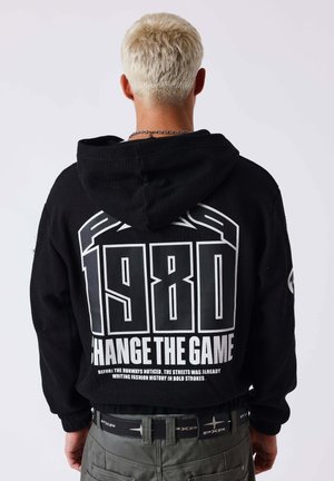 Person with short blond hair wears black hoodie with large white text "1980 Change the Game" on back against plain light background.