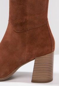 Bottine en daim marron avec talon bloc en bois superposé et surpiqûres visibles sur la partie supérieure de la chaussure.