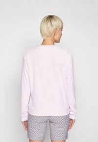 Απαλή ροζ ραμμένη μπλούζα με μακριά μανίκια, στρογγυλή λαιμόκοψη και πλεκτούς καρπούς, συνδυασμένη με καρό σορτς. Πίσω όψη, λεία υφή.