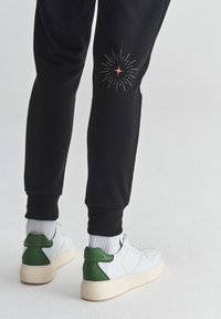 Piernas con pantalones de chándal negros que tienen un pequeño diseño de explosión de estrellas en la pantorrilla, calcetines blancos y zapatillas blancas con acentos verdes en el talón sobre un suelo claro.