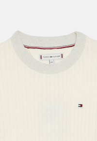 Kremfarget ribbestrikket genser med en lys grå, glitrende rund halsringning. Har en liten brodert logo i rødt, hvitt og blått.