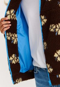 Hnědá fleecová bunda s květinovými vzory v béžové a modré barvě, s modrou podšívkou a zapínáním na zip.