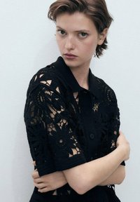 Black lace polo shirt featuring intricate floral patterns, short sleeves, and a buttoned collar. Soft texture with transparent details.