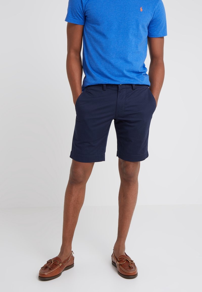 Homme portant un t-shirt bleu, un short bleu marine foncé et des chaussures bateau en cuir marron, debout les mains dans les poches.