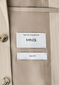 Intérieur de costume beige montrant deux étiquettes blanches : "THE SUIT COLLECTION MNG" et "SLIM FIT", avec deux boutons beiges visibles sur le côté gauche.