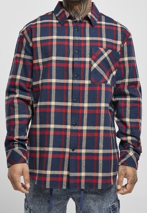 Man wearing long-sleeve blue, red, and beige plaid button-up shirt with chest pocket and tattoos visible on neck and hands.