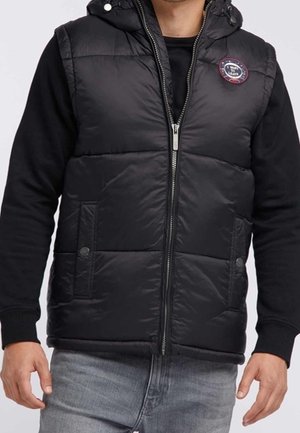 Homme portant un gilet rembourré noir avec deux poches à boutons-pression à l'avant, une fermeture éclair, une capuche et un écusson rond sur la poitrine, par-dessus une chemise noire à manches longues et un jean gris.