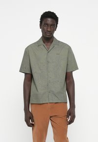 Short-sleeve button-up shirt in olive green with contour line pattern, featuring a fold-down collar and subtle logo detail on chest.