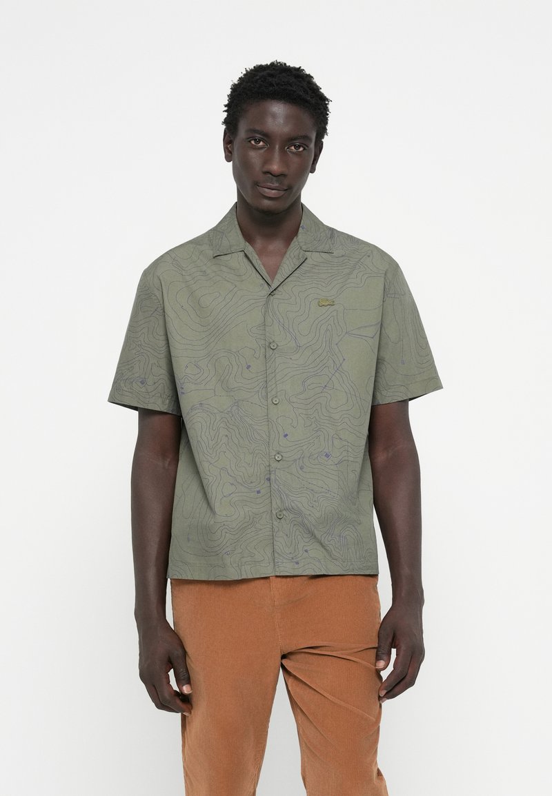 Short-sleeve button-up shirt in olive green with contour line pattern, featuring a fold-down collar and subtle logo detail on chest.