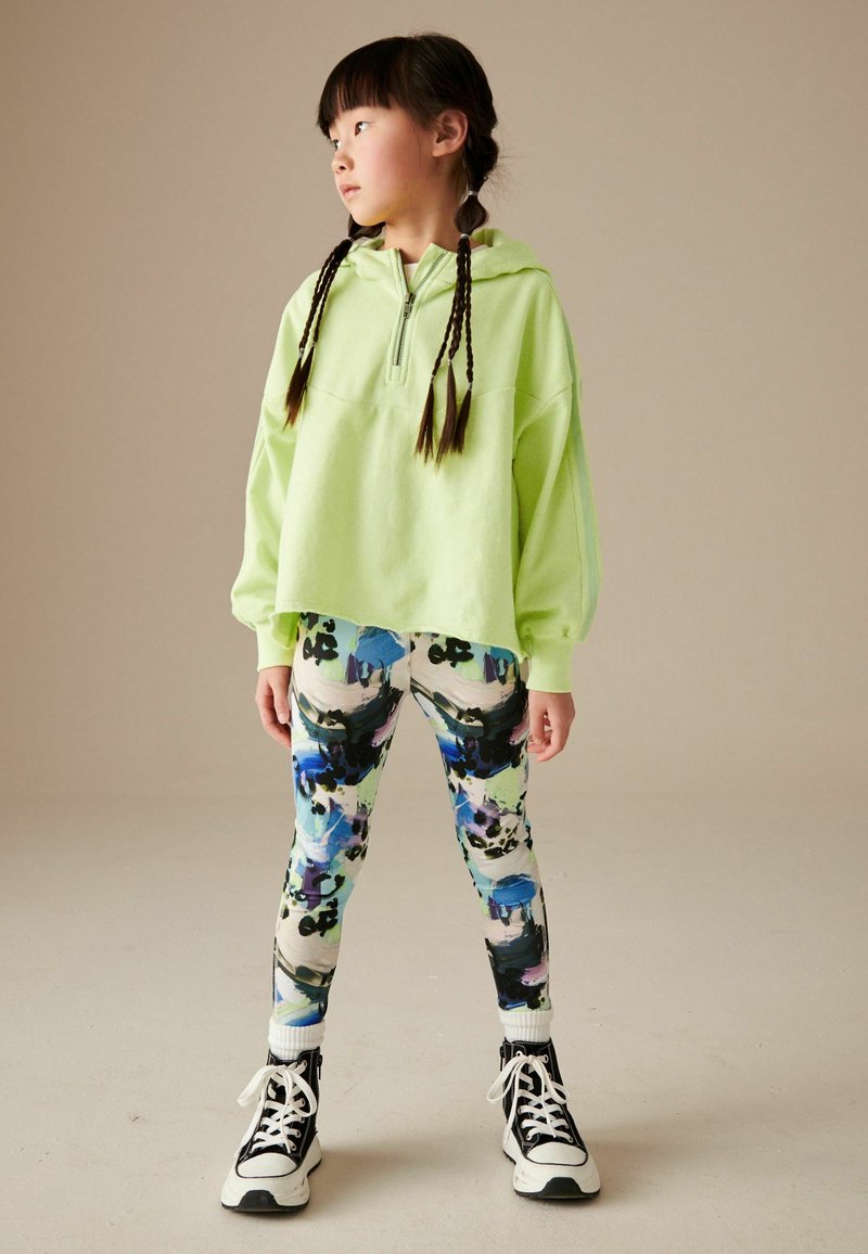 Ljusgrön huvtröja med frontdragkedja och puffärmar, kombinerad med leggings i färgglada abstrakta mönster och svarta sneakers.