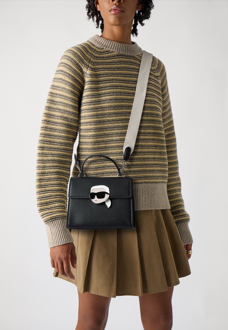 Borsa a tracolla in pelle nera con un design strutturato, dotata di una chiusura a fumetto, manico corto e una tracolla grigia regolabile. Persona che indossa un maglione a righe.