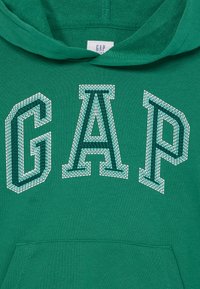 Zelená mikina s vreckom typu klokanka. Veľké logo "GAP" v bielej farbe s textúrou z zelených korálkov. Mäkko tkaný materiál, rebrovaná kapucňa a manžety.