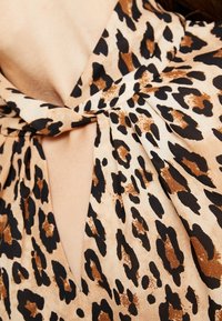 Animal print bluse med et brunt og sort leopardmønster på en beige stof, med en knude detalje ved halsen og plisseret design.