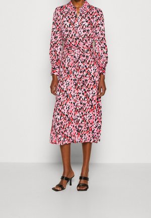 Mujer con un vestido rosa de manga larga hasta la rodilla con manchas abstractas rojas, negras y blancas, combinado con sandalias negras de tacón con tiras.