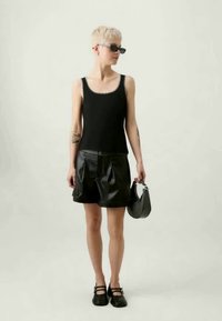 Top preto sem mangas com bordas em forma de onda, combinado com calções de pele pretos plissados e sapatos planos pretos. A segurar uma pequena mala preta.