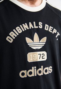 Melnā kokvilnas T-krekls ar krēmkrāsas apkakli, kurā ir uzraksts "ORIGINALS DEPT." un zelta Adidas logo, kā arī "1972" grafika zelta un melnā krāsā.