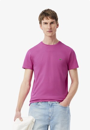 T-shirt rosa a maniche corte in cotone con un piccolo logo verde sul petto, abbinato a jeans di denim azzurro chiaro.