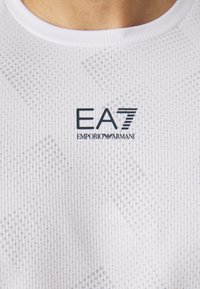 Bijela sportska majica s teksturom, okruglim izrezom i otisnutim logotipom "EA7 EMPORIO ARMANI" u tamnoplavoj boji sprijeda.