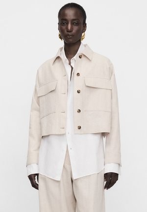 Donna di colore che indossa una giacca beige corta con bottoni su una camicia bianca e pantaloni beige coordinati, con orecchini grandi dorati visibili.