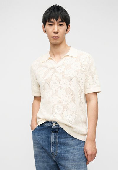 Πλεκτό κοντομάνικο πόλο σε κρεμ απόχρωση με μοτίβο λουλουδένιας δαντέλας, γιακά στο λαιμό και διακριτικές ριμπ λεπτομέρειες. Φορεμένο με μπλε τζιν.