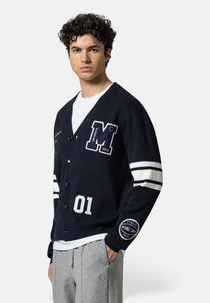 Young man with curly hair wearing a navy cardigan with white stripes and patches, paired with a white shirt and gray sweatpants.